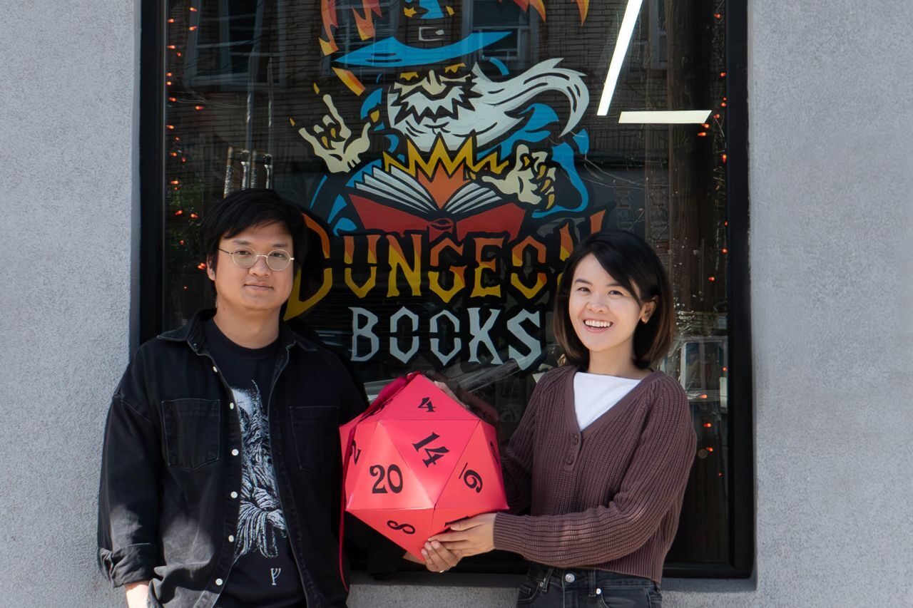 Panat and Carrie holding a giant red d20 in front of the Dungeon Books window, which is painted with the wizard logo