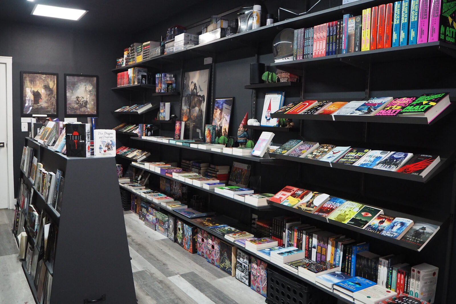 The long main wall of black shelves with books face out, framed fantasy prints above, a gray plank floor