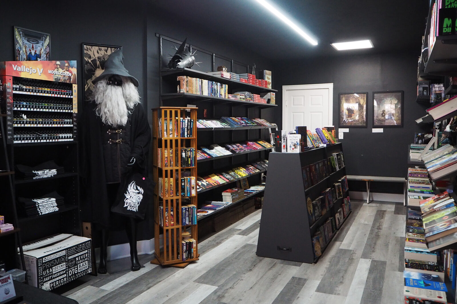 The RPG section: shelves of RPG books, a wooden spinner rack of paperbacks, a rack of Vallejo paints and a mannequin dressed as a gray-bearded wizard holding a Dungeon Books tote